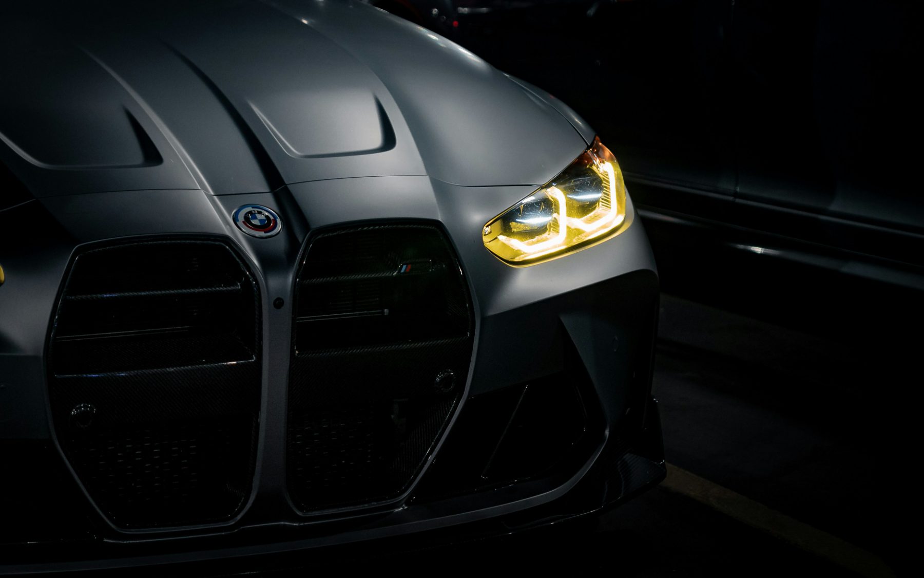 Matte-grey BMW M with one headlight illuminated in a dark garage
